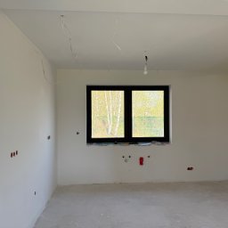Sobex Team - Wnętrze pokoju po szpachlowaniu ścian i sufitu, widoczne gładkie powierzchnie, okno z czarną ramą i wystające kable elektryczne. Przygotowanie do malowania.