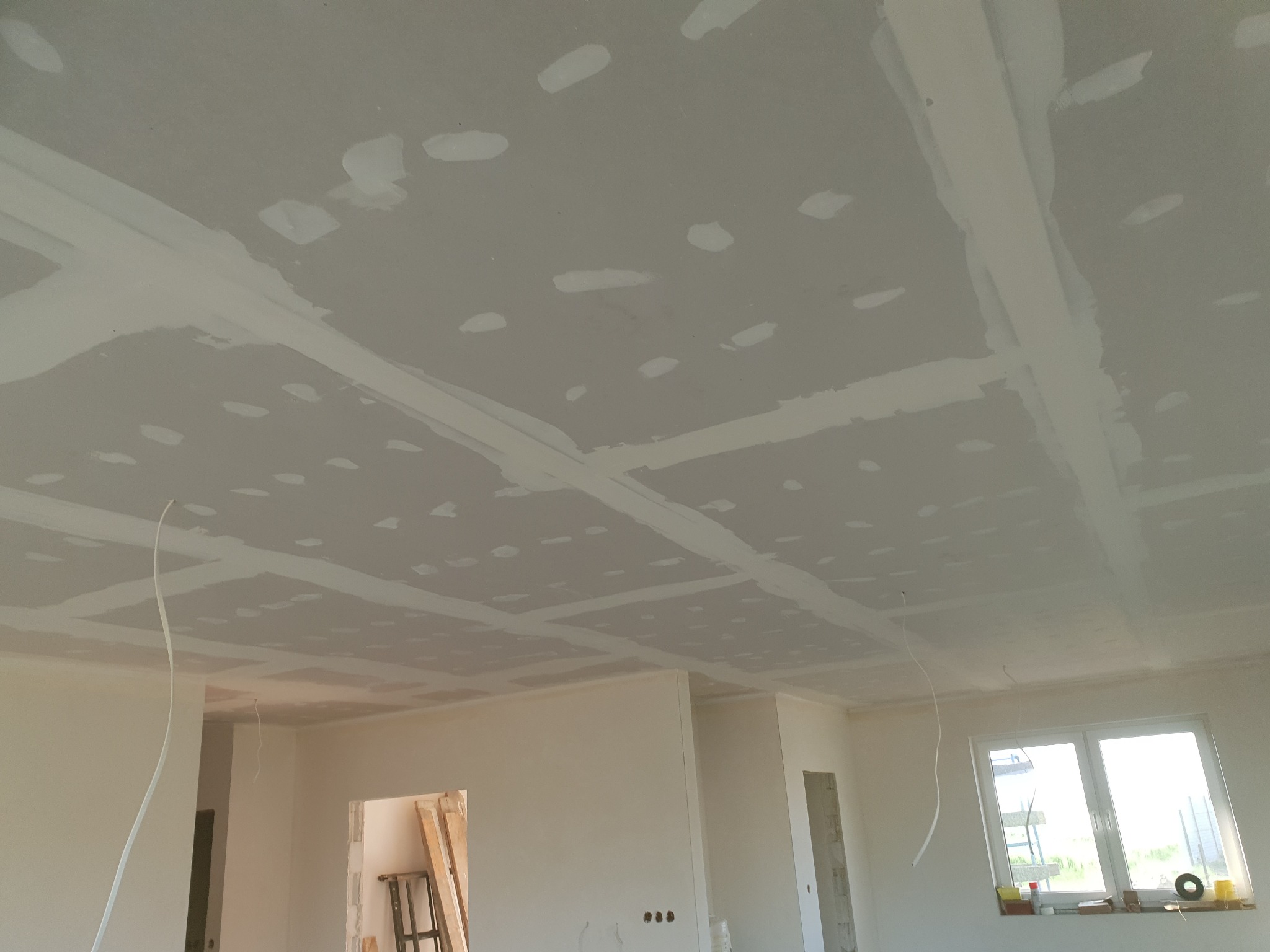Sufit z płyt gipsowo-kartonowych po szpachlowaniu, widoczne łączenia płyt i miejsca po wkrętach. W tle fragment ściany i okno z parapetem.