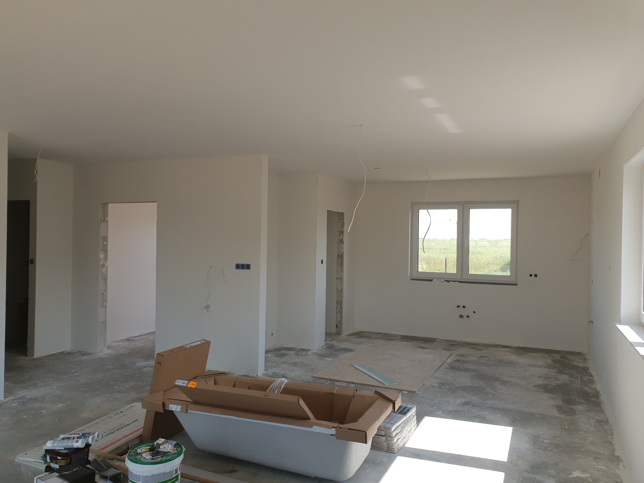 Wnętrze w trakcie remontu: białe ściany po szpachlowaniu, okno z widokiem na zewnątrz, kartony i materiały budowlane na podłodze. Przygotowanie do dalszych prac wykończeniowych.