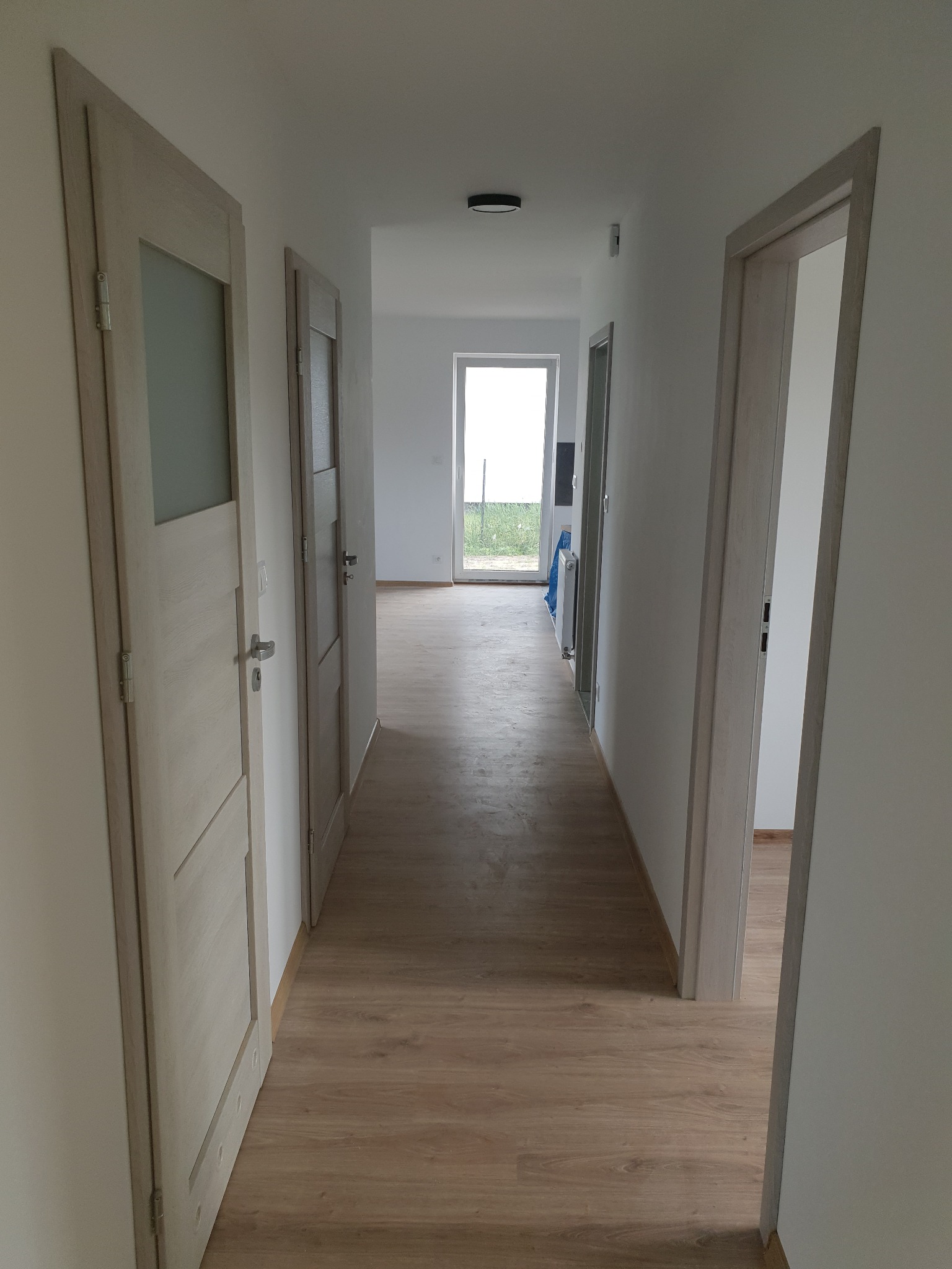 Jasne, przestronne wnętrze z trzema parami drzwi w odcieniach beżu i szarości, prowadzące do kolejnych pomieszczeń. Podłoga laminowana w ciepłym odcieniu drewna.