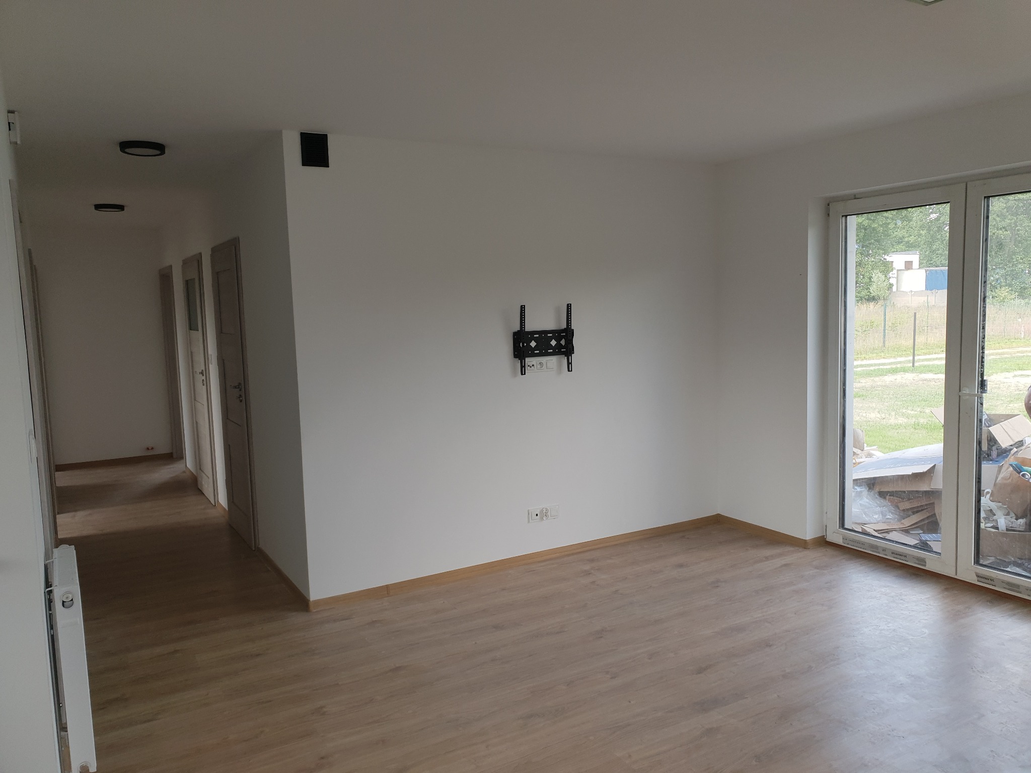 Jasne, puste wnętrze po remoncie z drewnianą podłogą, białymi ścianami i zamontowanym uchwytem na telewizor. Widoczne okno balkonowe i drzwi w głębi.