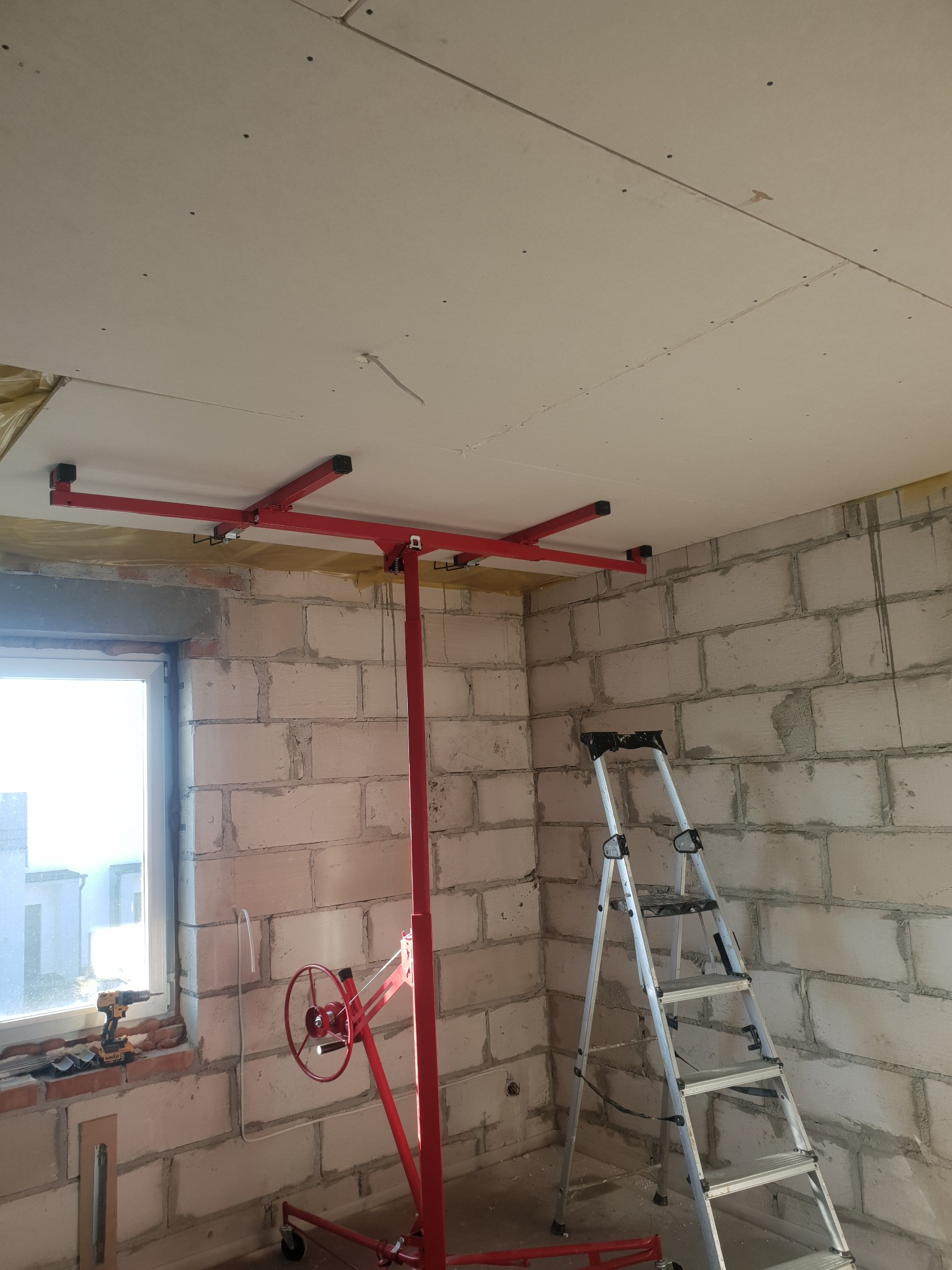 Montaż płyty na suficie za pomocą podnośnika. Widoczna drabina, surowe ściany z pustaków i narzędzia przy oknie. Etap prac wykończeniowych w nowym budynku.
