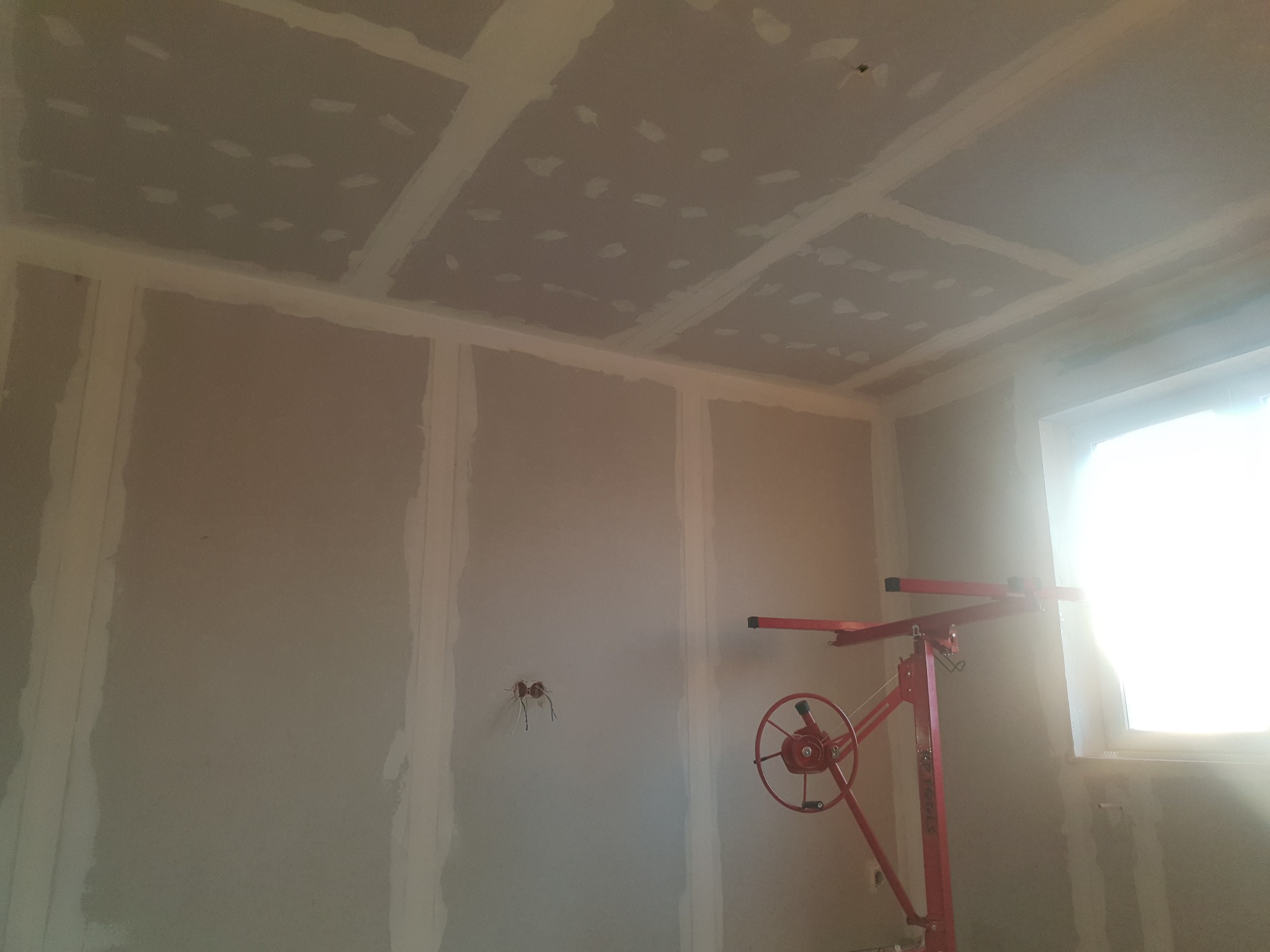 Wnętrze w trakcie montażu płyt gipsowo-kartonowych na ścianach i suficie, widoczny podnośnik do płyt, częściowo wykończone łączenia, surowe gniazdka elektryczne.