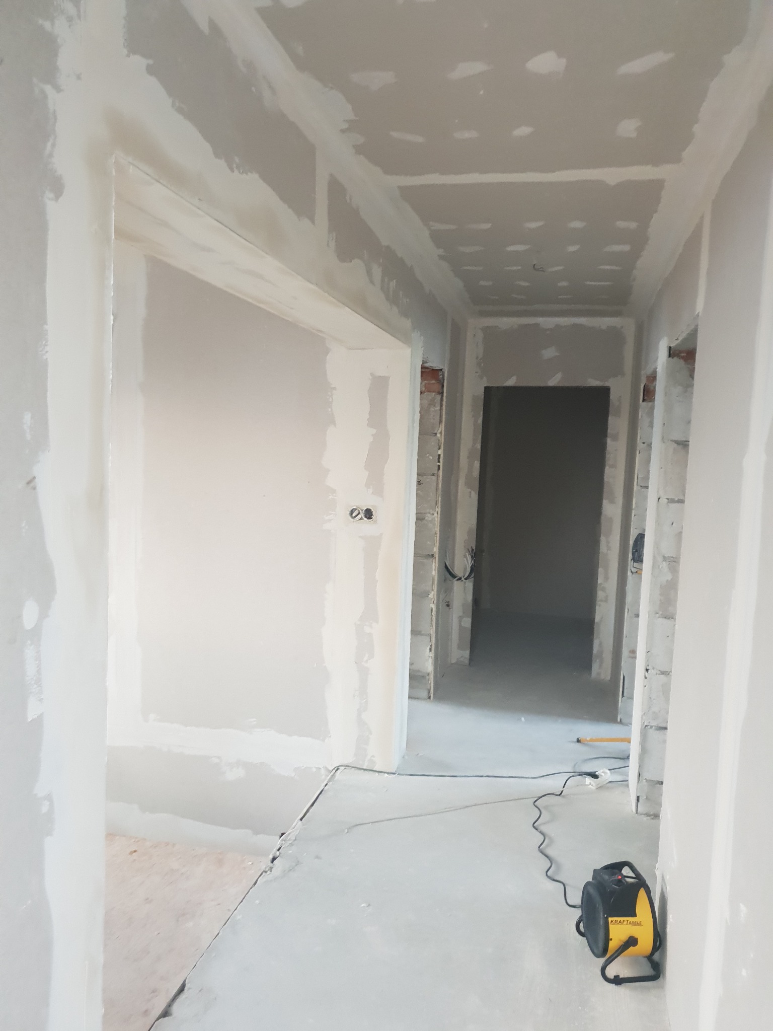 Korytarz w trakcie szpachlowania i montażu płyt karton-gips. Widoczne ściany i sufit pokryte szpachlą, surowy stan wykończenia, grzejnik budowlany na podłodze.
