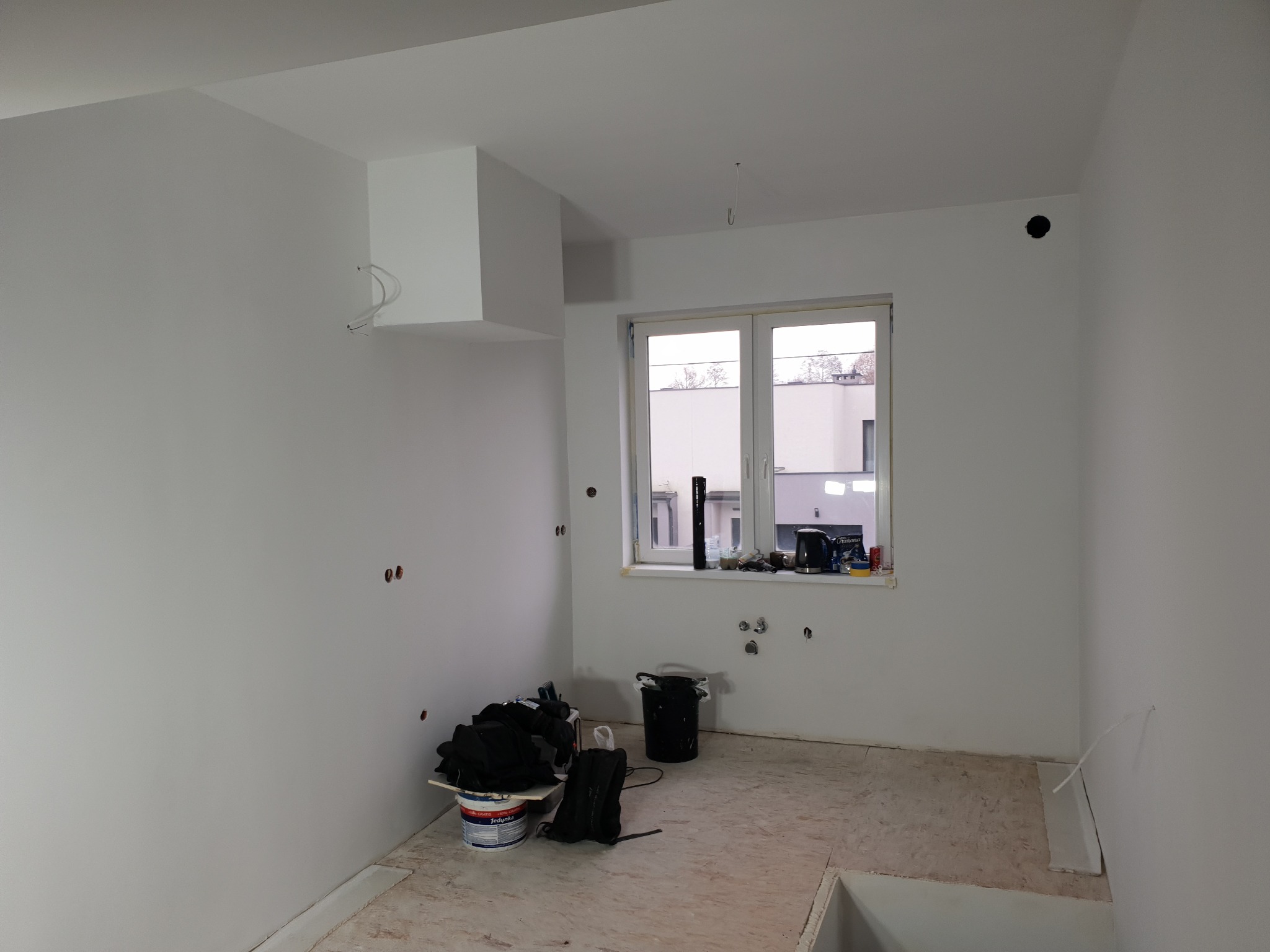 Wnętrze w trakcie remontu: białe ściany po szpachlowaniu, okno z widokiem na zewnątrz, widoczne instalacje elektryczne i hydrauliczne, przygotowane materiały budowlane.