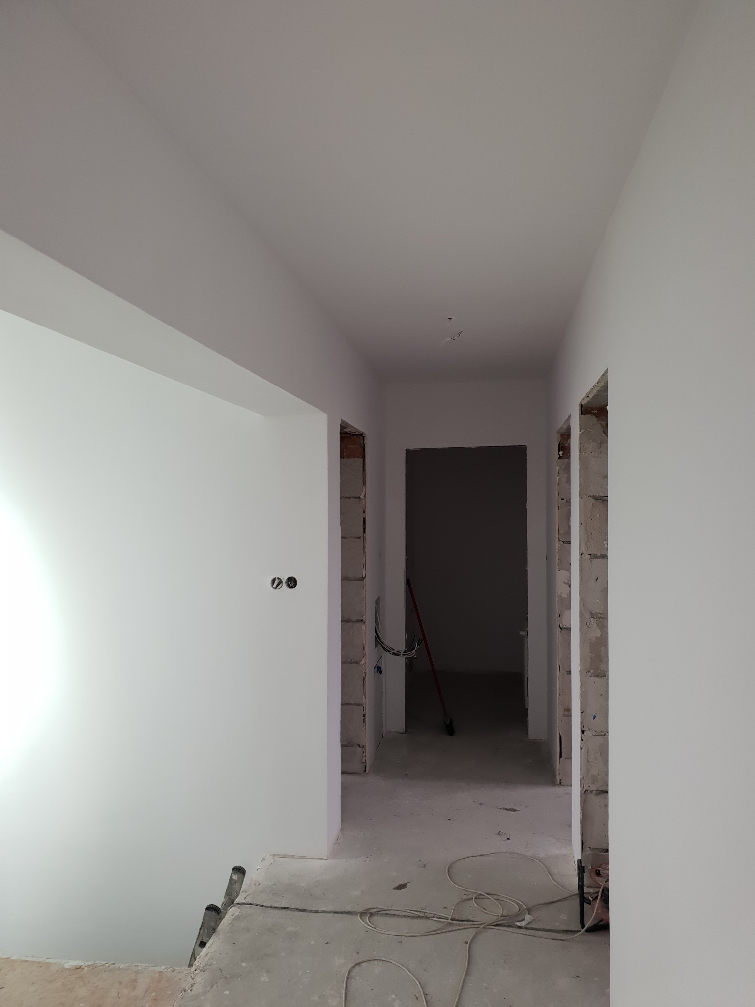 Wnętrze po szpachlowaniu: gładkie, białe ściany w korytarzu z widocznymi puszkami elektrycznymi i surowymi ościeżami. Podłoga betonowa, widoczne narzędzia.