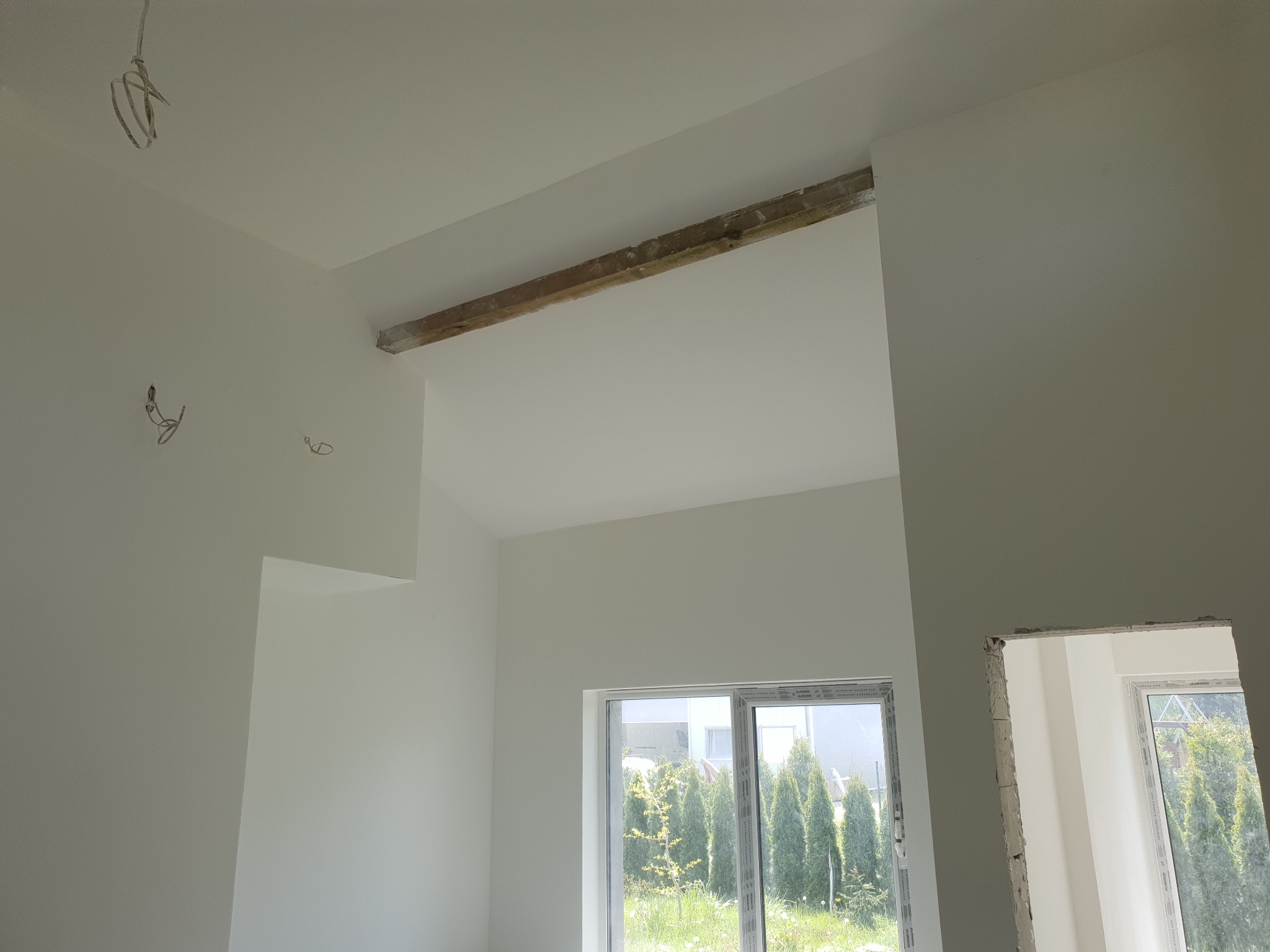 Wnętrze po remoncie: świeżo pomalowane ściany na biało, widoczne okna z widokiem na zieleń, drewniana belka pod sufitem i wystające kable elektryczne.