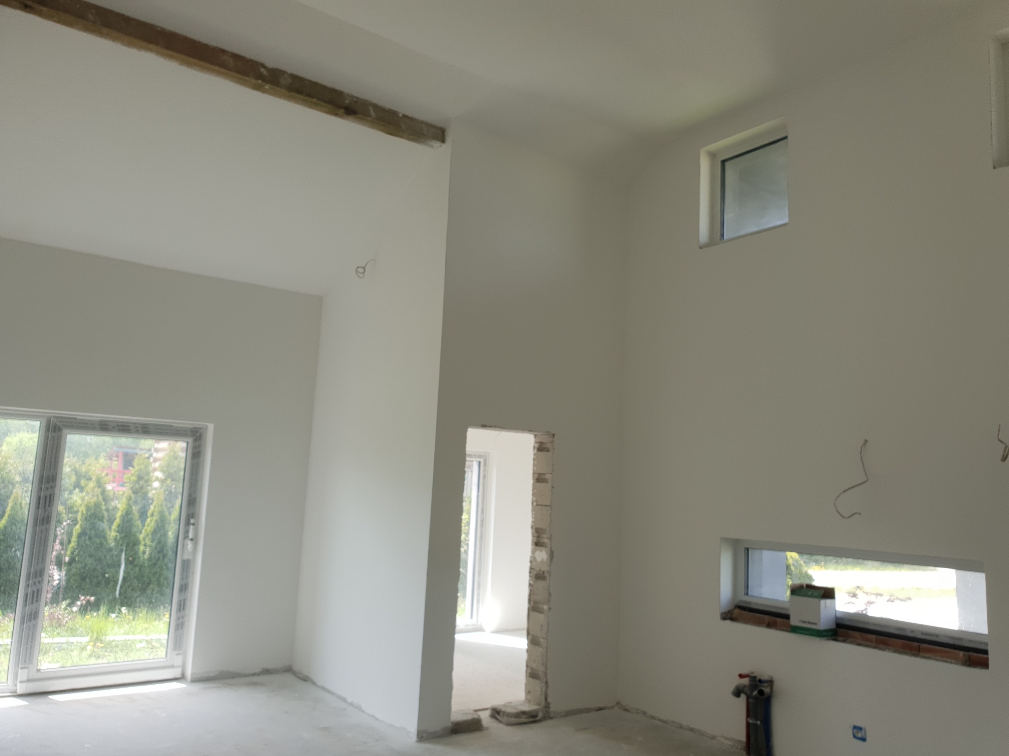 Wnętrze po szpachlowaniu: gładkie, białe ściany z oknami o różnych kształtach i rozmiarach. Widoczne ślady prac wykończeniowych i surowy drewniany element pod sufitem.