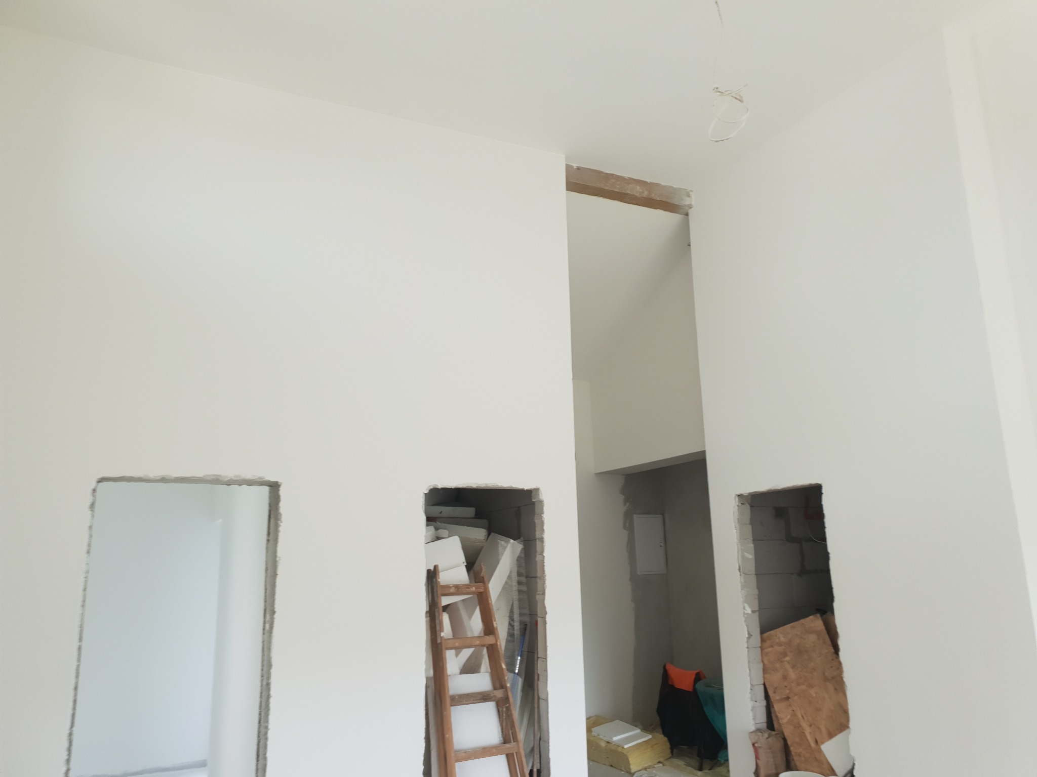 Białe ściany po szpachlowaniu z trzema otworami, w jednym drabina i materiały budowlane. Widoczne niedoskonałości w otworach.