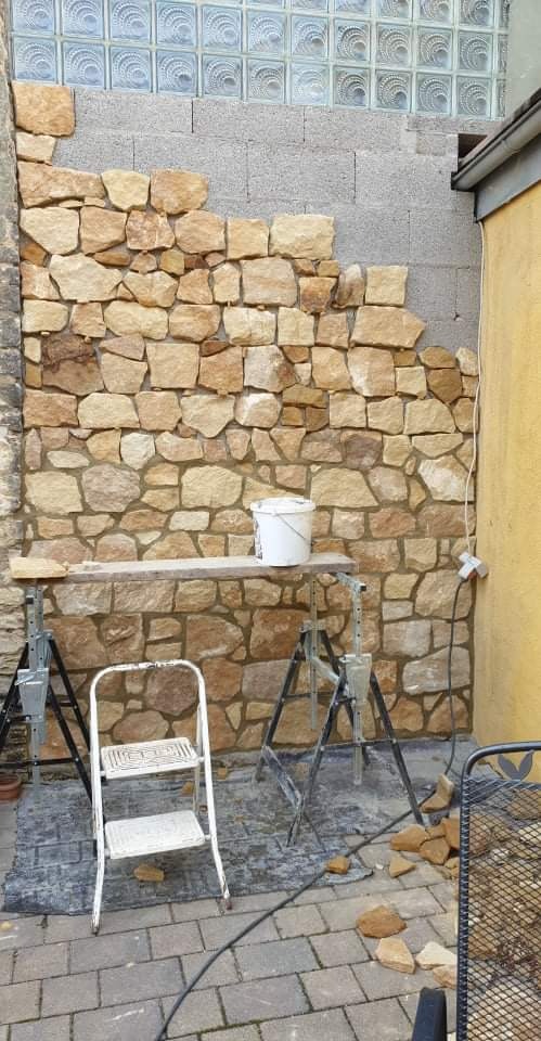 Fragment ściany zewnętrznej w trakcie obkładania nieregularnym kamieniem, widoczne rusztowanie, drabina i wiadro z zaprawą, w tle ściana z luksferów.