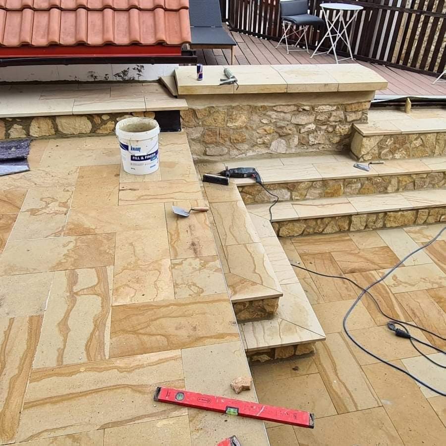 Układanie piaskowych płytek na tarasie i schodach, widoczna poziomica, wiadro z zaprawą i narzędzia glazurnicze.