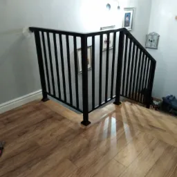 Czarne metalowe balustrady schodowe z pionowymi szczeblami, zamontowane na drewnopodobnych panelach podłogowych, z parą szczypiec leżących na podłodze w lewym dolnym rogu.