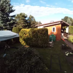 Widok ogrodu z równo przyciętym żywopłotem z tuj, drewnianą altaną z numerem 490, zielonym pojemnikiem na deszczówkę w kształcie żółwia, oraz metalową drabinką na trawniku.