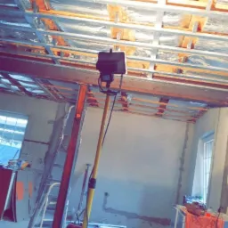 Wnętrze w trakcie prac wykończeniowych: widoczna konstrukcja sufitu podwieszanego z izolacją, drabina oparta o ścianę, lampa budowlana na statywie i plastikowe pojemniki na podłodze.