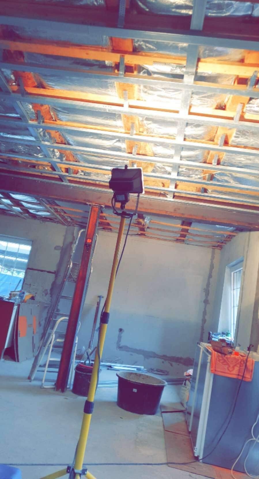 Wnętrze w trakcie prac wykończeniowych: widoczna konstrukcja sufitu podwieszanego z izolacją, drabina oparta o ścianę, lampa budowlana na statywie i plastikowe pojemniki na podłodze.
