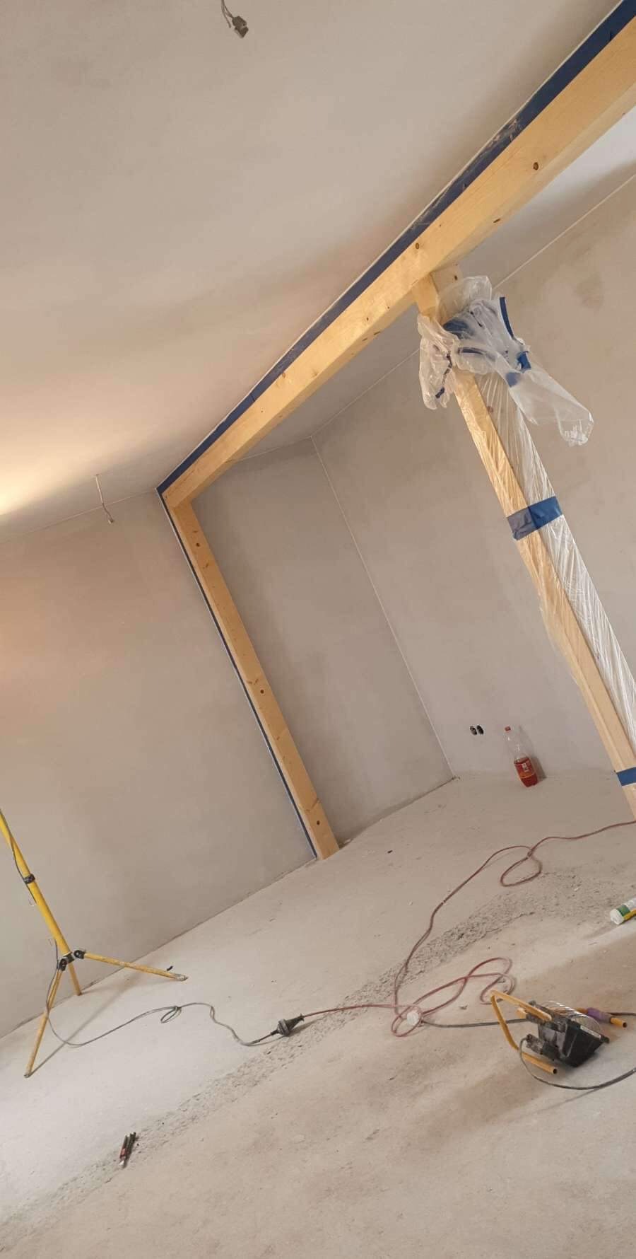Wnętrze w trakcie prac: drewniana konstrukcja oznaczona taśmą malarską zabezpieczająca otwór, plastikowa folia ochronna, lampa budowlana na trójnogu oraz narzędzia i przewody na podłodze.