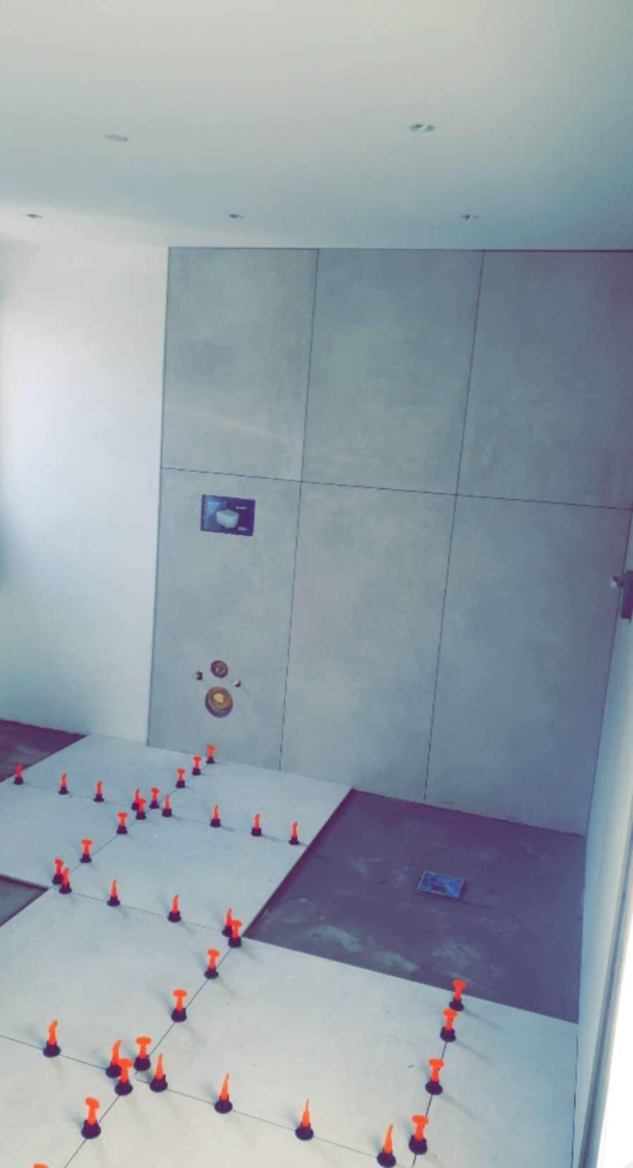 Łazienka w trakcie kładzenia płytek podłogowych i ściennych w szarym odcieniu, z widocznymi pomarańczowymi elementami poziomującymi.