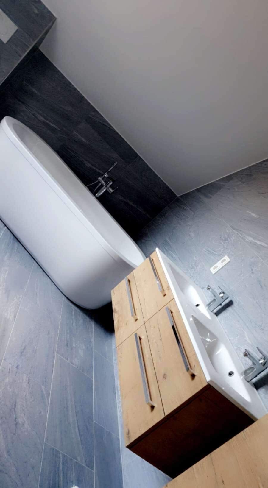 Nowoczesna łazienka z wolnostojącą wanną, drewnianą szafką pod umywalką i szarymi płytkami imitującymi kamień na ścianach.