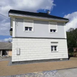 Dwukondygnacyjny dom z szarą elewacją górnej części i białą elewacją imitującą cegłę w dolnej części, z roletami zewnętrznymi w oknach, anteną satelitarną na dachu i częściowo ułożoną kostką...