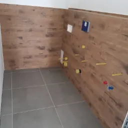 Łazienka w trakcie remontu: szare płytki podłogowe, drewnopodobne panele ścienne, widoczne instalacje hydrauliczne z żółtymi rurami i zaworami.