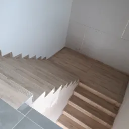 Montaż paneli na schodach z wykończeniem płytami kartonowo-gipsowymi na ścianie. Widok z góry na drewnopodobne panele i szare płytki podłogowe.