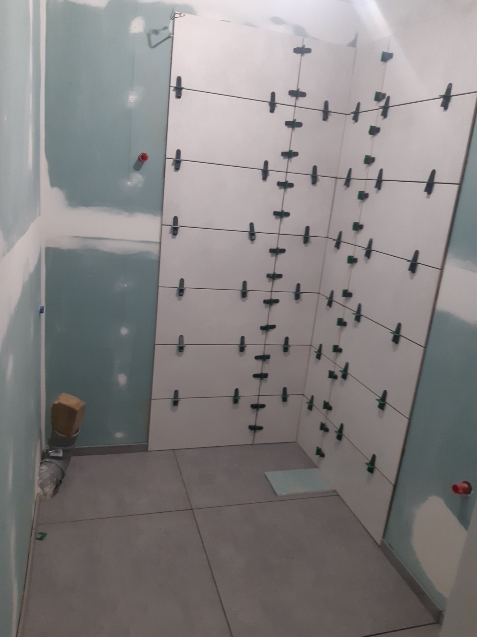 Białe płytki ścienne z czarnymi krzyżykami dystansowymi w rogu łazienki, szare płytki podłogowe, ściany przygotowane do malowania. Etap prac wykończeniowych.