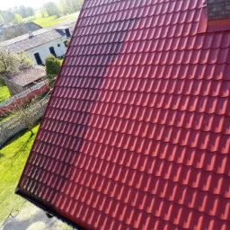 Czerwony dach z blachodachówki, widoczne zabrudzenia i zacienienia, w tle zabudowania gospodarcze i zieleń.