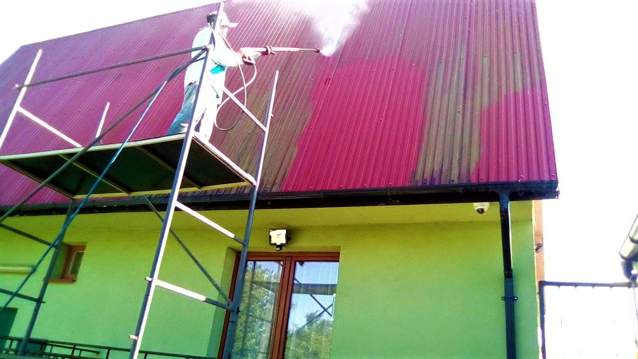 Pracownik w kombinezonie ochronnym na rusztowaniu czyści metalowy, bordowy dach z użyciem myjki ciśnieniowej, widoczna różnica w czystości dachu przed i po umyciu.