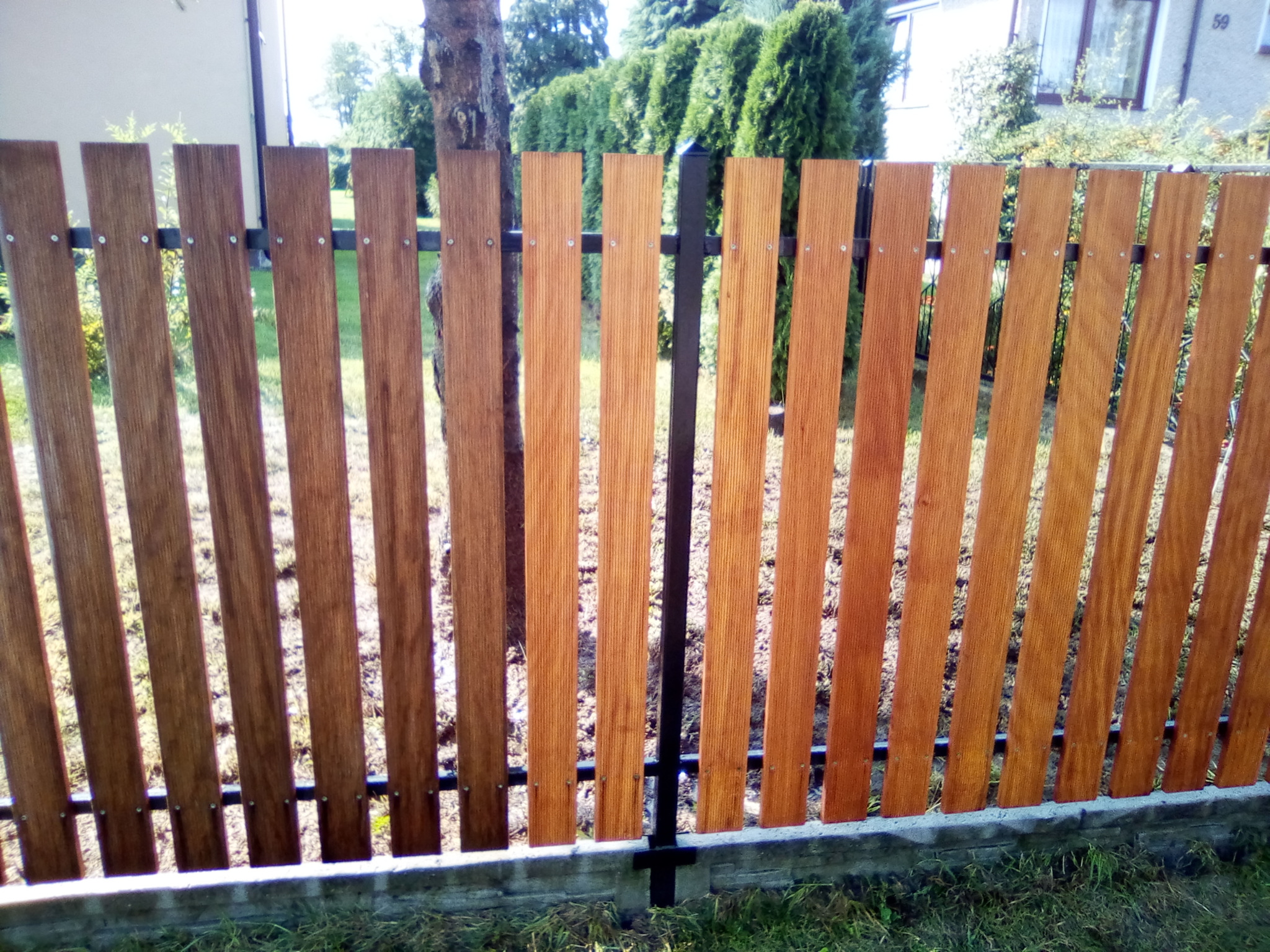 Drewniany płot z pionowych desek o ciepłym, brązowym odcieniu, przymocowanych do czarnej, metalowej ramy, z widocznym betonowym fundamentem i zielenią w tle.