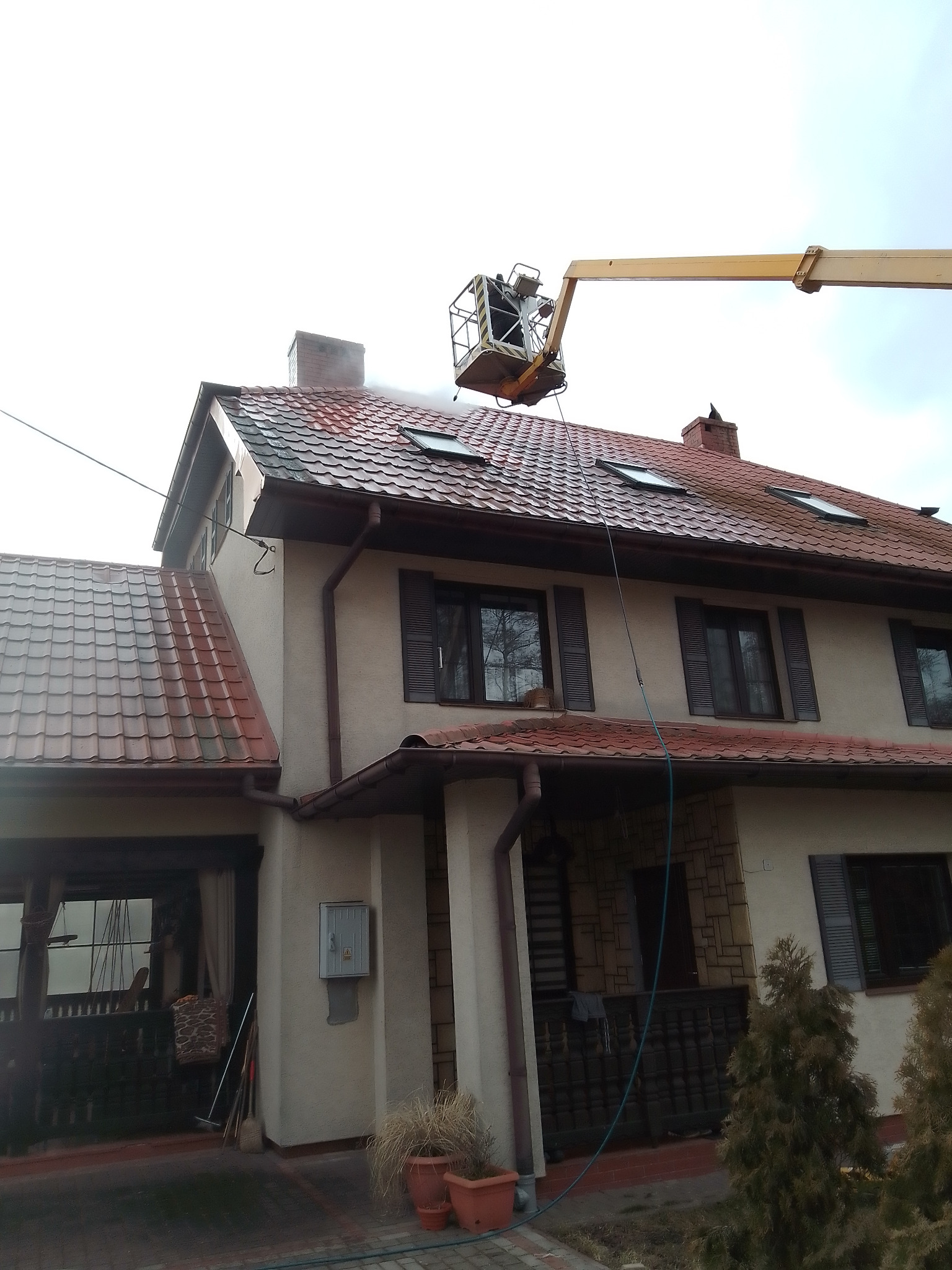 Dwupiętrowy dom z czerwonym dachem czyszczony z podnośnika koszowego, woda spryskuje dach, widoczne okna dachowe i rynny.