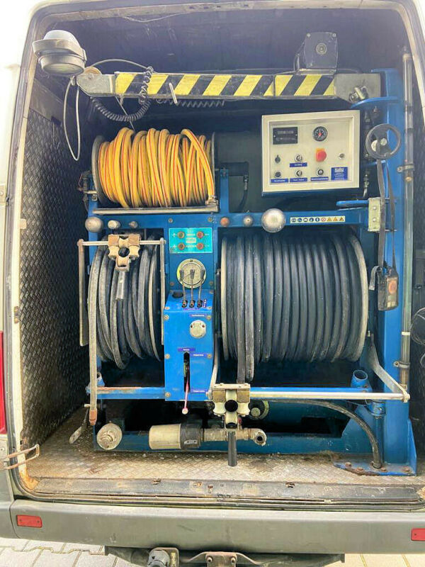 Wnętrze furgonetki z niebieskim urządzeniem do czyszczenia, z nawiniętymi wężami żółtym i czarnym, panel sterowania z przyciskami i miernikami, widoczne elementy hydrauliczne i elektryczne.