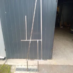 Konstrukcja metalowa, prawdopodobnie stojak, spawana z profili kwadratowych, umieszczona przed garażem z blachy trapezowej.