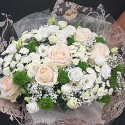 Bukiet z kremowych róż, białych eustom i drobnych białych chryzantem, owinięty w ozdobny papier z motywem kwiatowym w odcieniach różu i szarości, widoczny fragment dłoni.