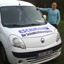 Biały samochód dostawczy firmy Eschmann, specjalizującej się w izolacji akustycznej, z pracownikiem opartym o maskę na tle zabudowań mieszkalnych.