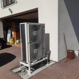 Zewnętrzna jednostka grzewcza z dwoma wentylatorami zamontowana na metalowym stelażu przy ścianie budynku z widocznym garażem i kostką brukową.