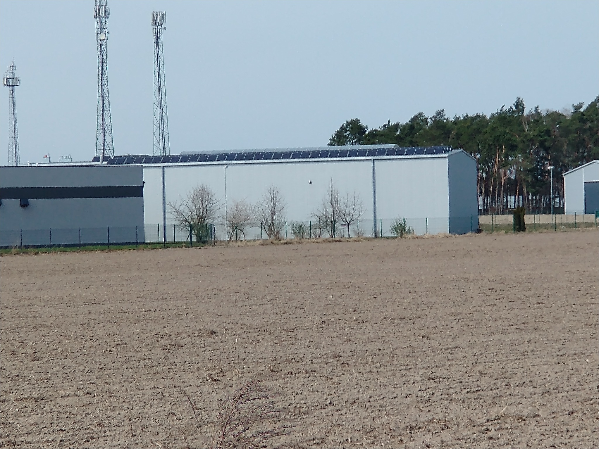 Długi budynek przemysłowy z panelami słonecznymi na dachu, widziany z pola uprawnego, w tle maszty telekomunikacyjne.