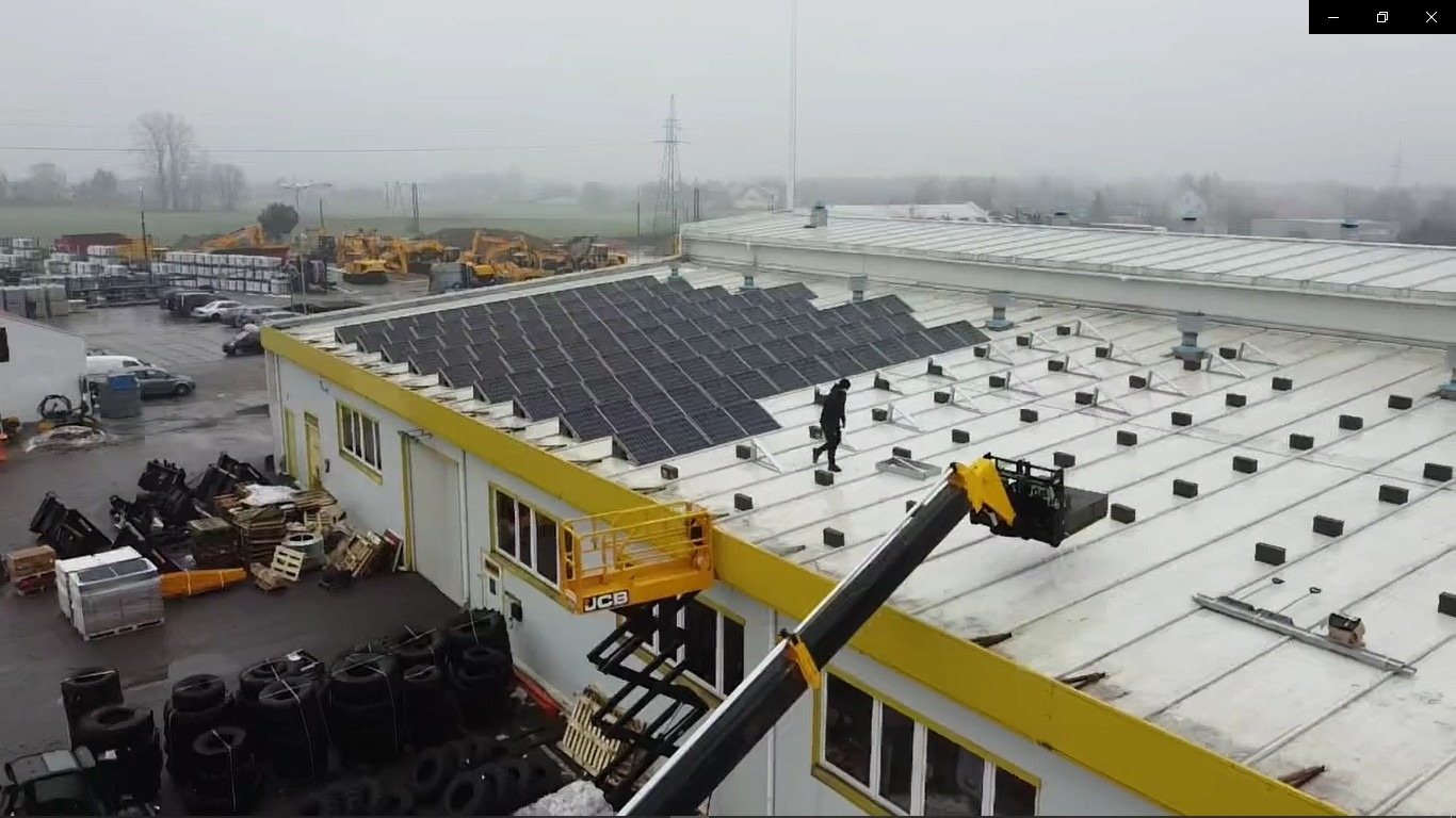 Montaż paneli słonecznych na dachu hali przemysłowej z użyciem podnośnika koszowego JCB; w tle plac z maszynami budowlanymi w pochmurny dzień.