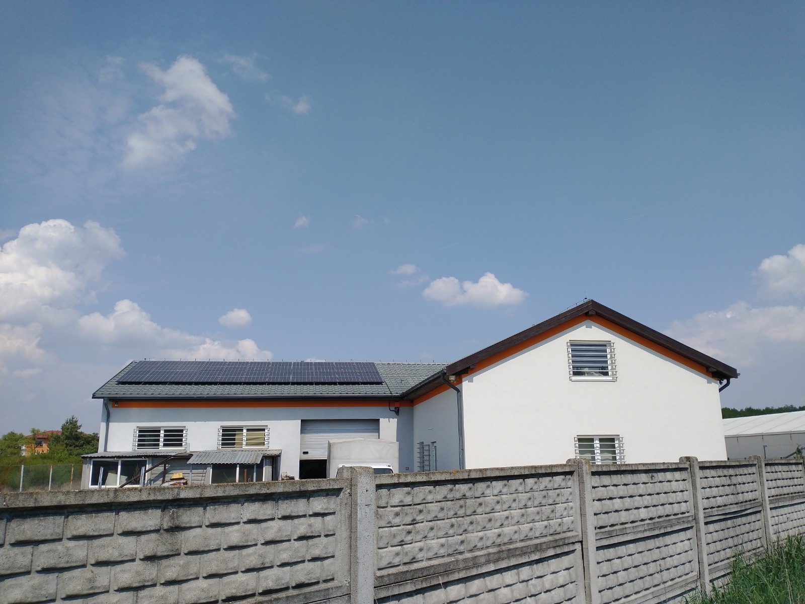 Budynek z panelami słonecznymi na dachu, widziany zza betonowego płotu w słoneczny dzień.