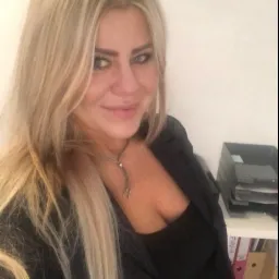 Portret kobiety z blond włosami, ubranej w czarną marynarkę, stojącej w biurze z półką na dokumenty w tle.