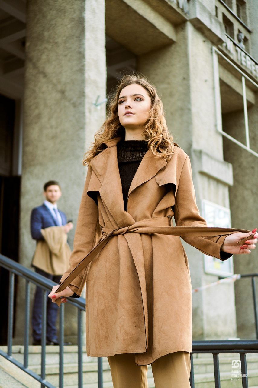 Kobieta w brązowym płaszczu na tle budynku, mężczyzna w garniturze w tle, ujęcie z dołu.