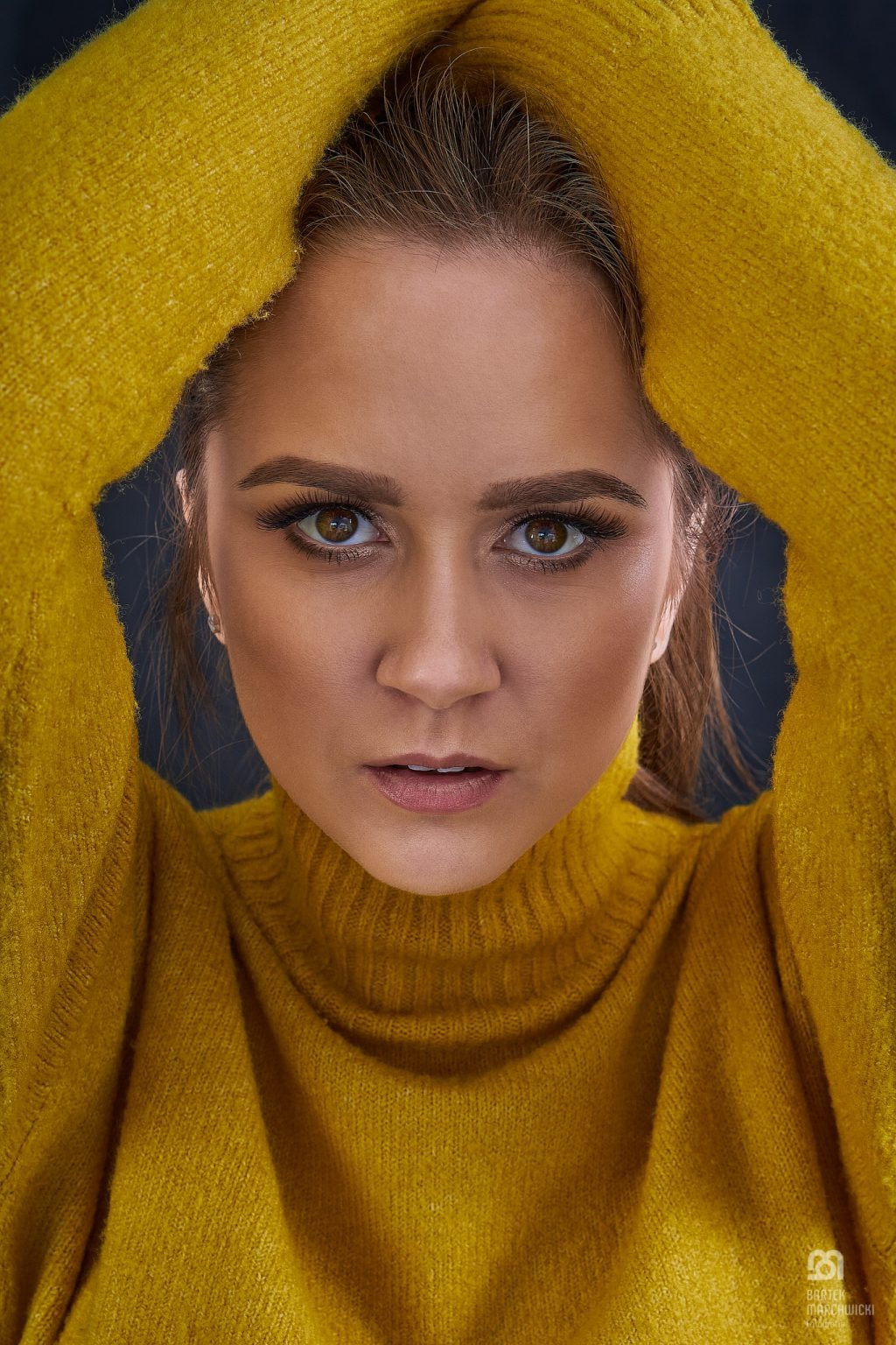 Portret kobiety w żółtym swetrze z golfem, ręce uniesione nad głową, intensywne spojrzenie w obiektyw.