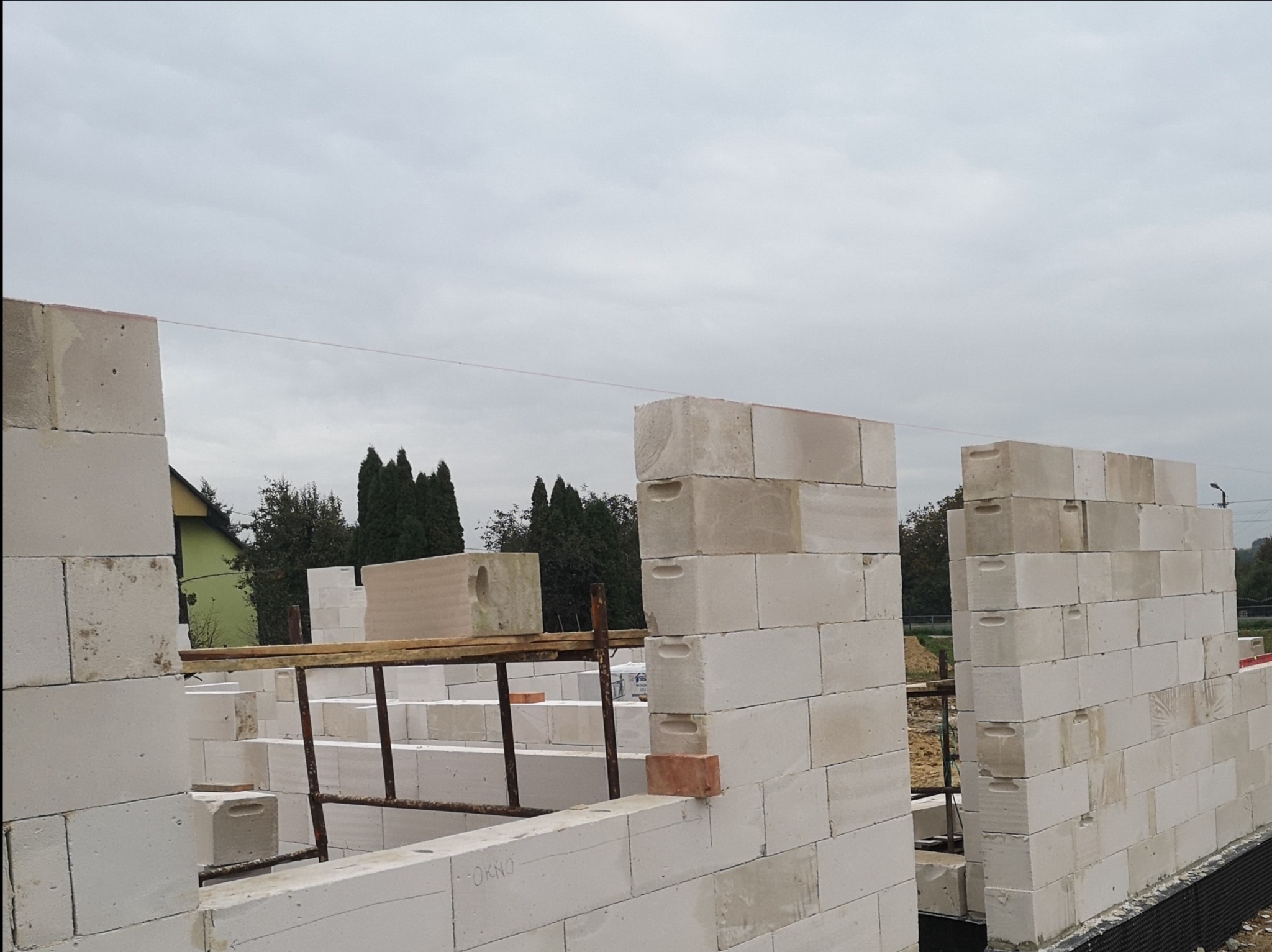 Wznoszone ściany z białych bloczków betonowych na budowie domu, widoczne rusztowania i pojedyncza czerwona cegła wmurowana w ścianę, pochmurne niebo w tle.