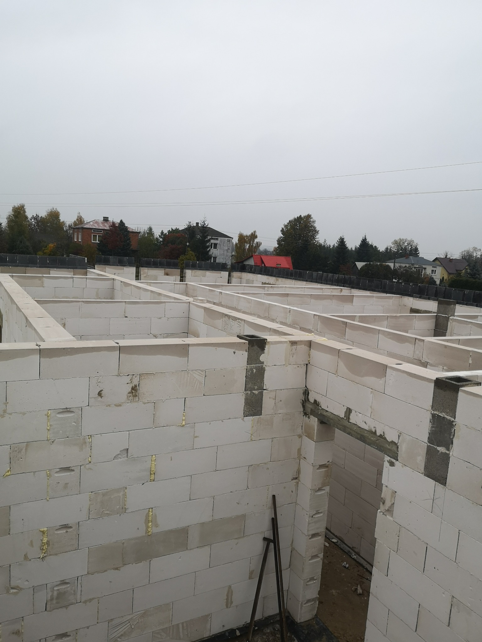 Wznoszone ściany fundamentowe z bloczków betonowych, widoczne spoiny wypełnione pianką montażową, w tle zabudowa jednorodzinna i pochmurne niebo.