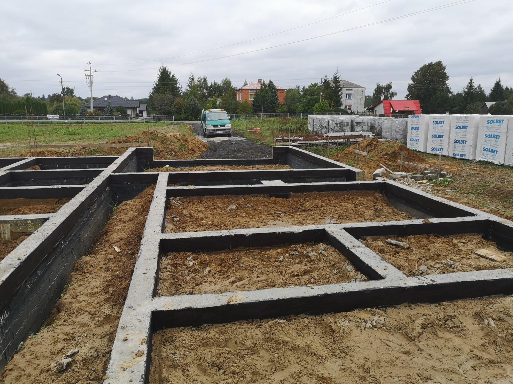 Zalane betonem szalunki fundamentowe na działce budowlanej, widoczne bloczki Solbet i zielony bus w tle.