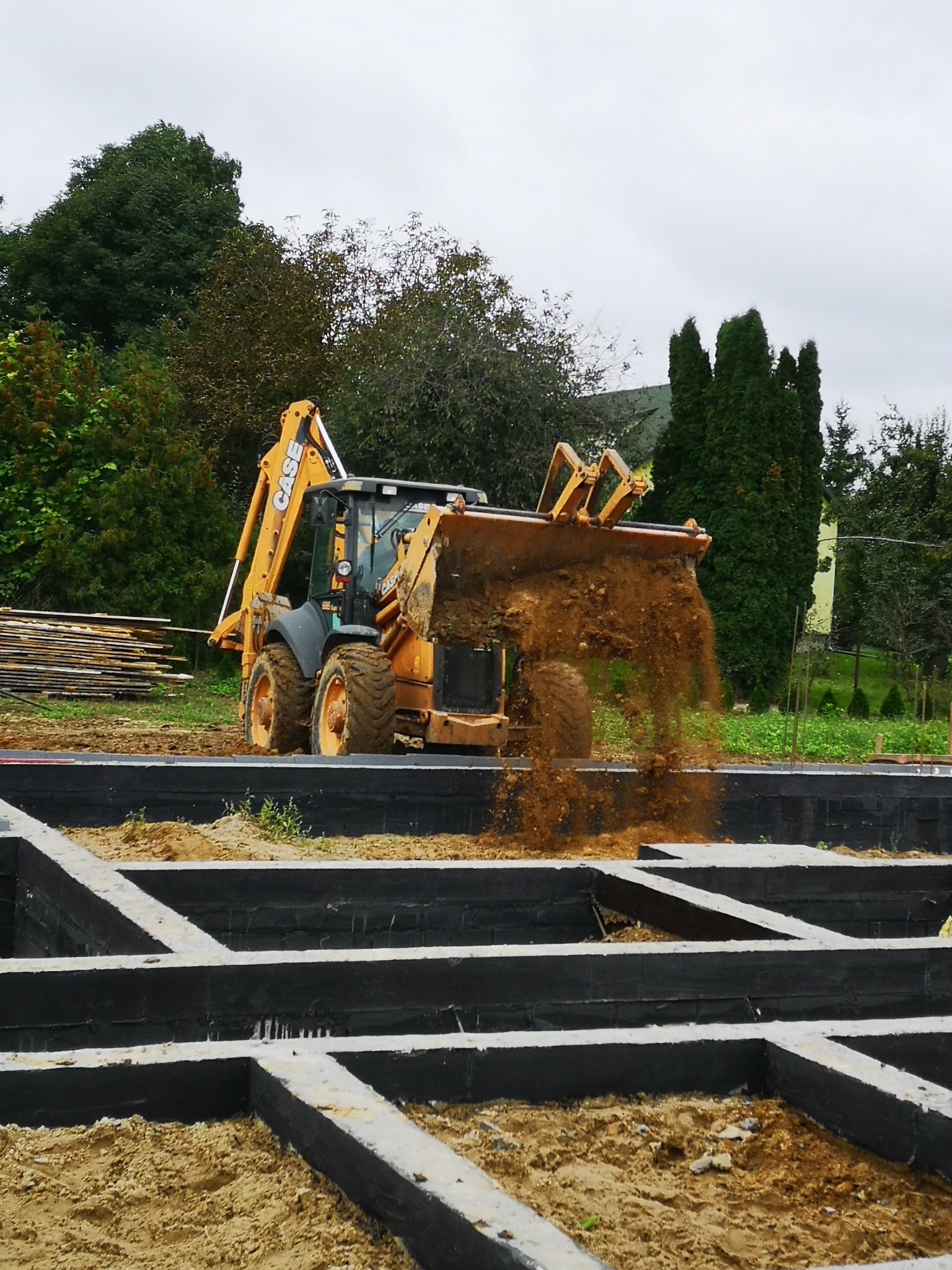 Żółto-czarna koparko-ładowarka CASE zasypuje piaskiem zarys fundamentów domu jednorodzinnego, widoczne deski i zieleń w tle.