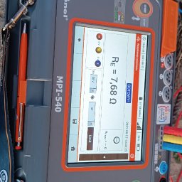 Szymon Drązewski PUH Rzemieślnik - Miernik Sonel MPI-540 z wyświetlonym wynikiem pomiaru rezystancji uziemienia Re=7.68Ω, przewody pomiarowe wpięte, tło z trawy i ziemi.