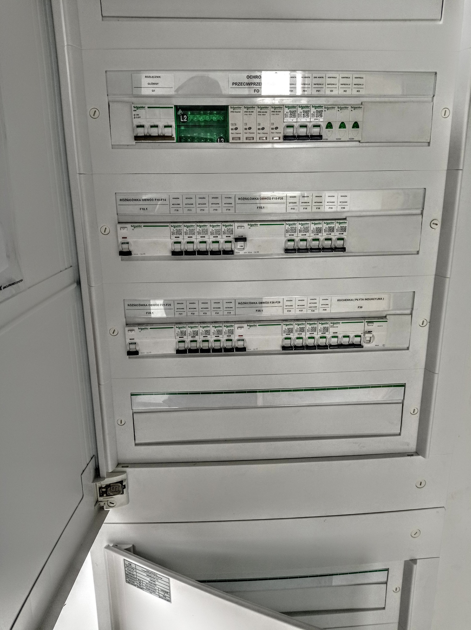 Rozdzielnia elektryczna z widocznymi bezpiecznikami i wyłącznikami różnicowoprądowymi marki Schneider Electric, oświetlona z góry, z otwartymi drzwiczkami dolnego segmentu.
