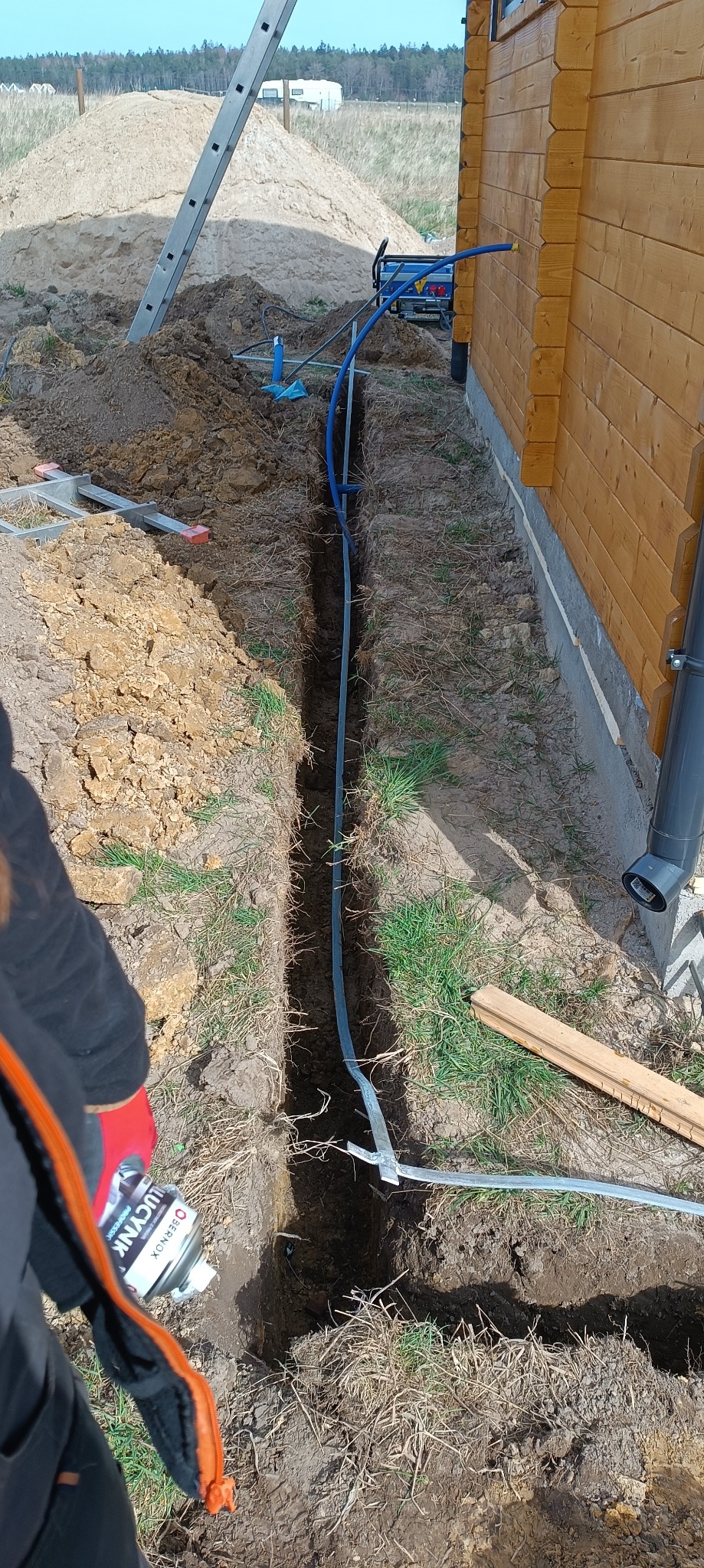Wykop pod przyłącze do drewnianego domu szkieletowego. W wykopie ułożone rury. Widoczna drabina, agregat i osoba z preparatem do zabezpieczenia instalacji.
