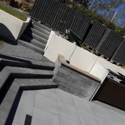 Nowoczesne schody z kostki brukowej w Złotowie, prowadzące na wyższy poziom ogrodu z widocznymi elementami drenażu i paletami z kostką w tle.