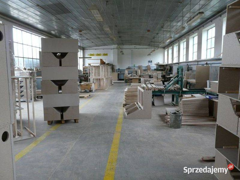 Wnętrze hali produkcyjnej z ułożonymi w stos elementami meblowymi z surowego drewna, widoczne maszyny stolarskie i materiały.
